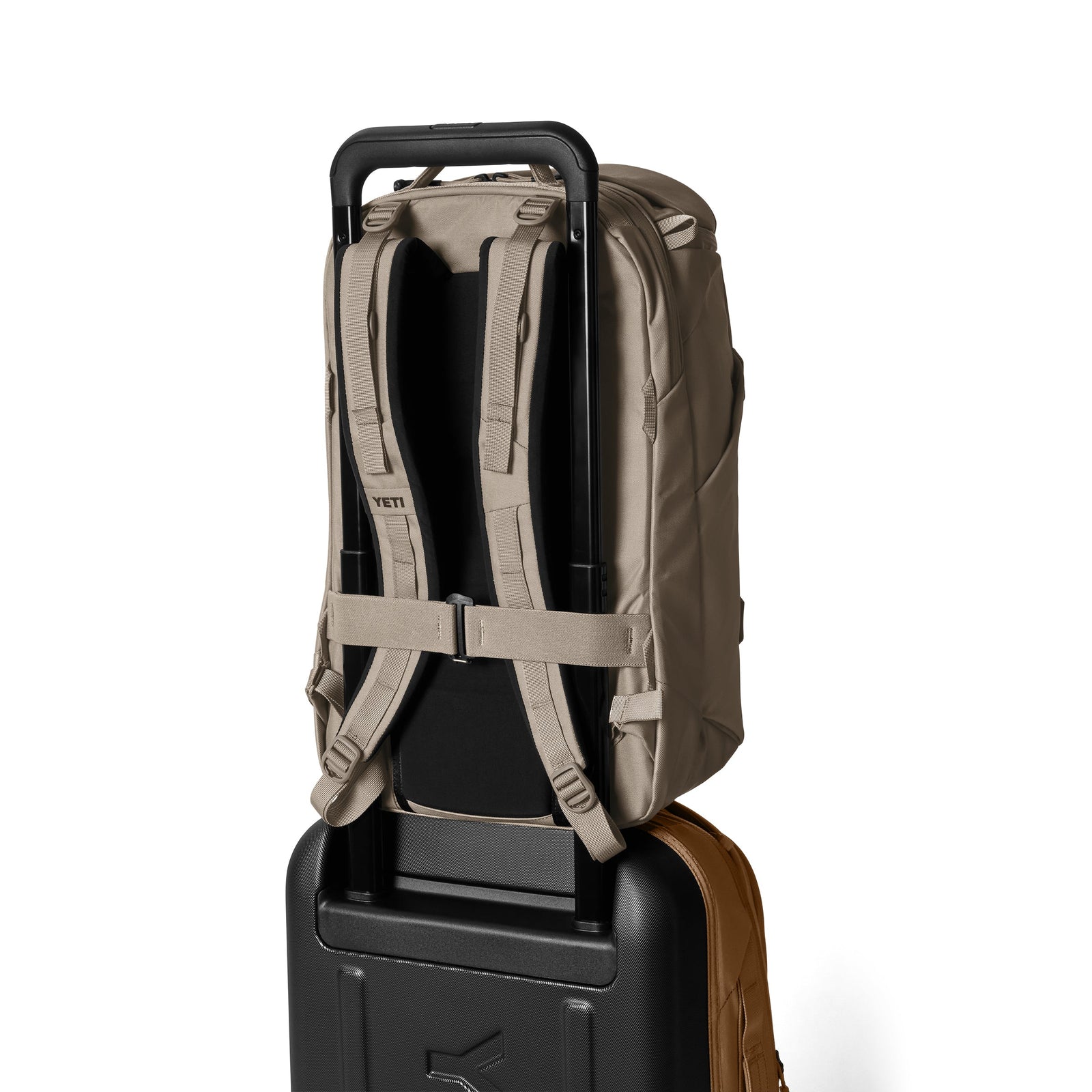 Der YETI® Rucksack Ranchero™ 27L in Cape Dark Taupe, mit sichtbaren Schultergurten, ist sicher am Griff eines schwarzen Rollkoffers befestigt. Beide stehen aufrecht vor einem weißen Hintergrund.