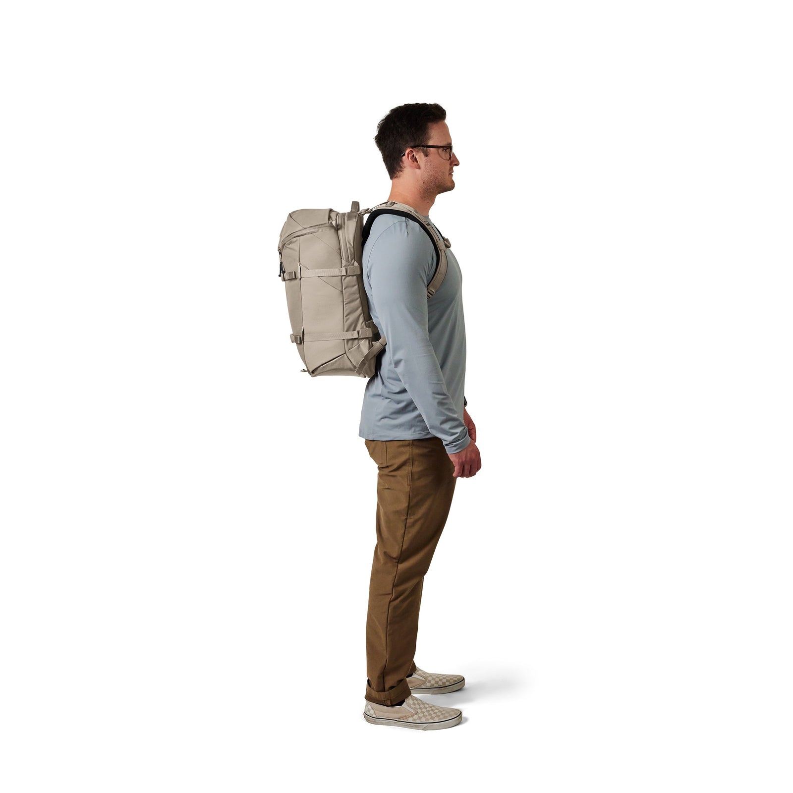 Mann im Profil vor weißem Hintergrund, trägt eine Brille, ein hellblaues langärmeliges Hemd, braune Hosen, Turnschuhe und hat einen YETI® Rucksack Ranchero™ 27L - Cape Dark Taupe dabei.