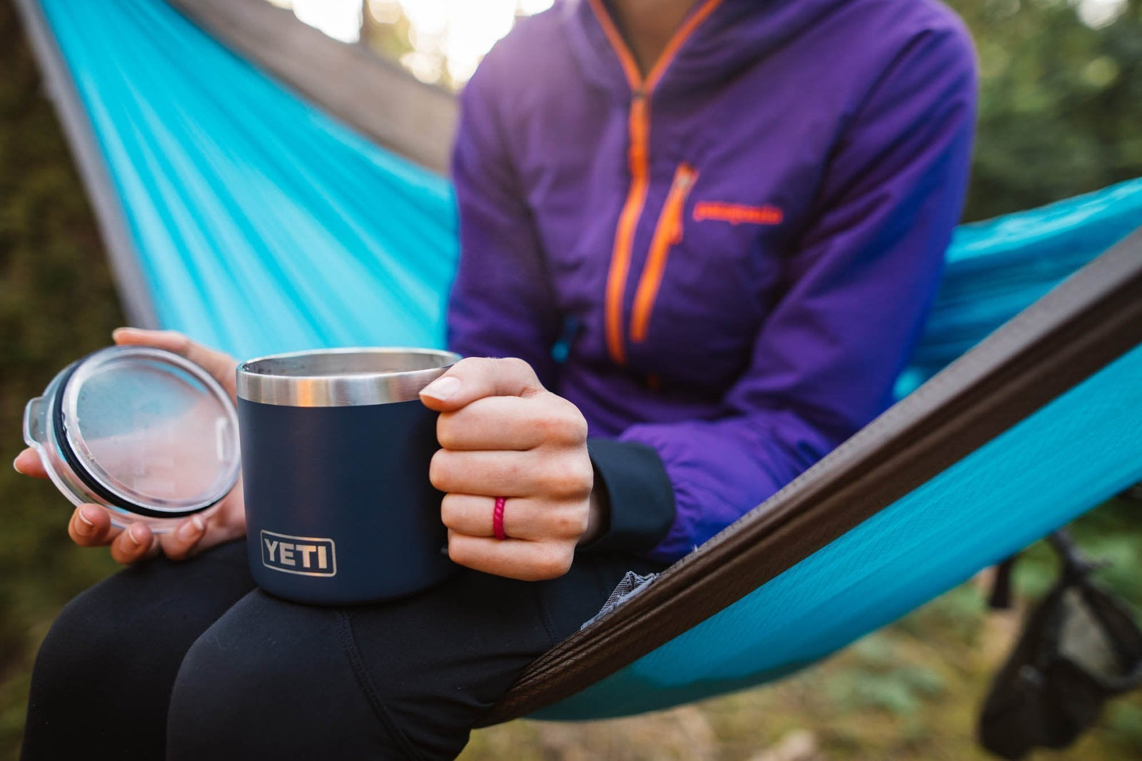 Eine Person in einer lilafarbenen Jacke entspannt sich auf einer blauen Hängematte und hält einen YETI® Tasse Rambler 14 Oz - Black in der einen und den Deckel in der anderen Hand, wodurch eine gemütliche Campingatmosphäre entsteht.
