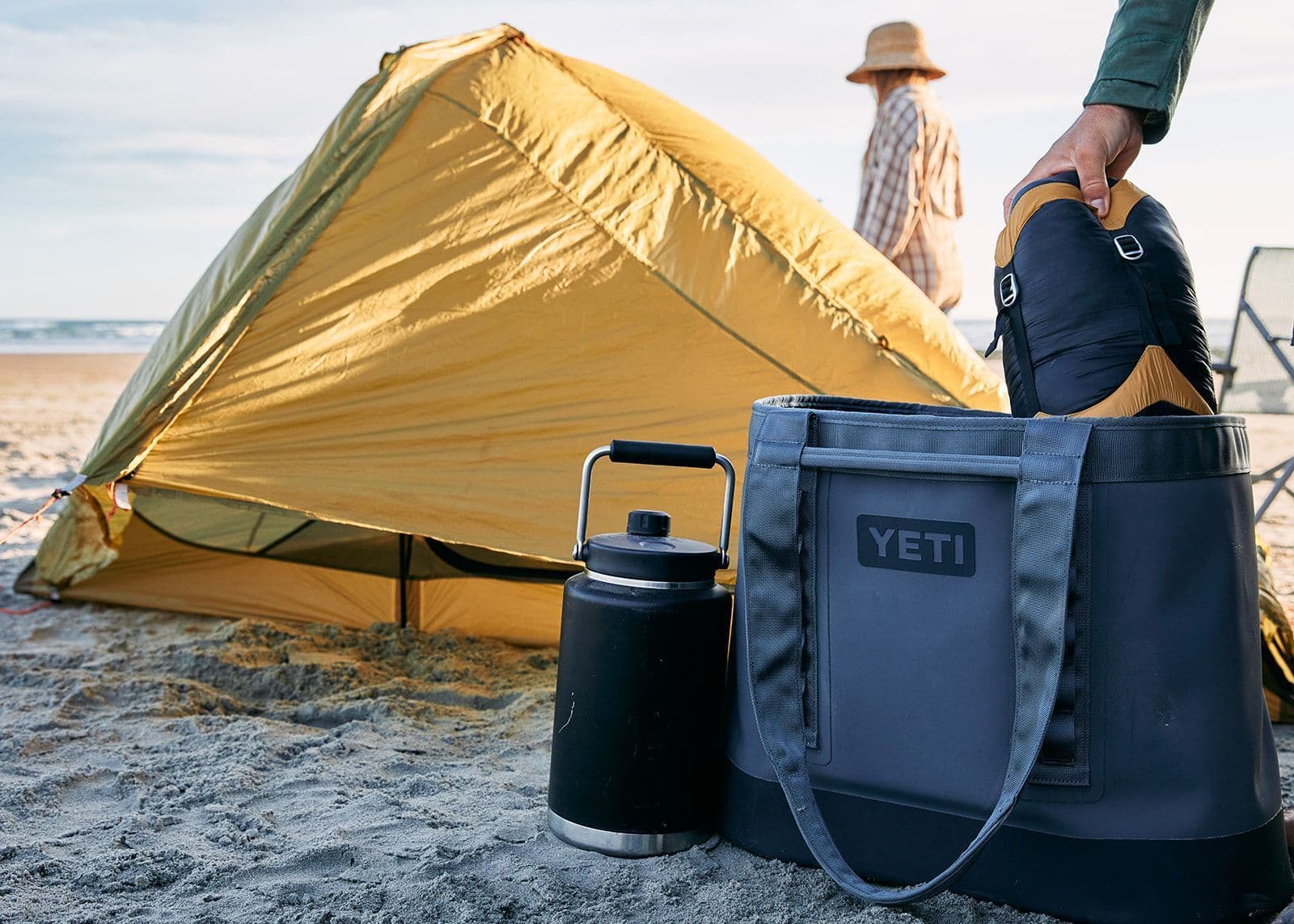 Eine Person packt einen Schlafsack in eine YETI® Tragetasche Camino® Carryall 50 - Storm Grey auf sandigem Boden neben einem gelben Zelt und einer Wasserflasche; diese robuste, wasserdichte Tasche von YETI ist perfekt für Outdoor-Abenteuer.