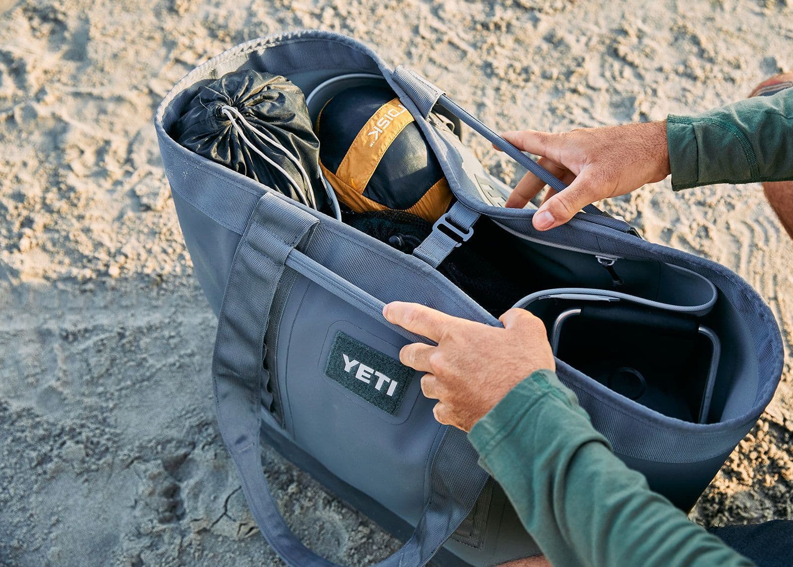 Eine Person öffnet eine YETI® Tragetasche Camino® Carryall 50 - Storm Grey, die mit Campingutensilien wie einem zusammengerollten Zelt oder Schlafsack gefüllt ist, während sie auf sandigem Boden sitzt.