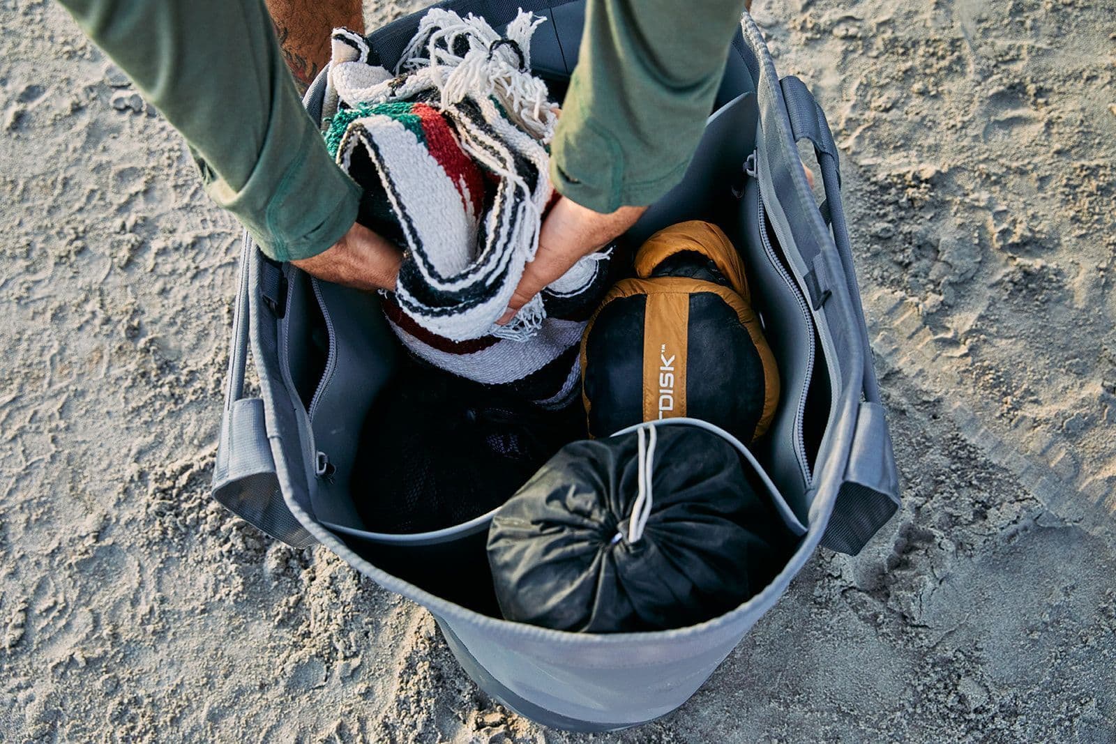 Auf sandigem Boden stehend, packt eine Person eine YETI® Tragetasche Camino® Carryall 50 - Storm Grey mit einer gestreiften Decke und Campingausrüstung.