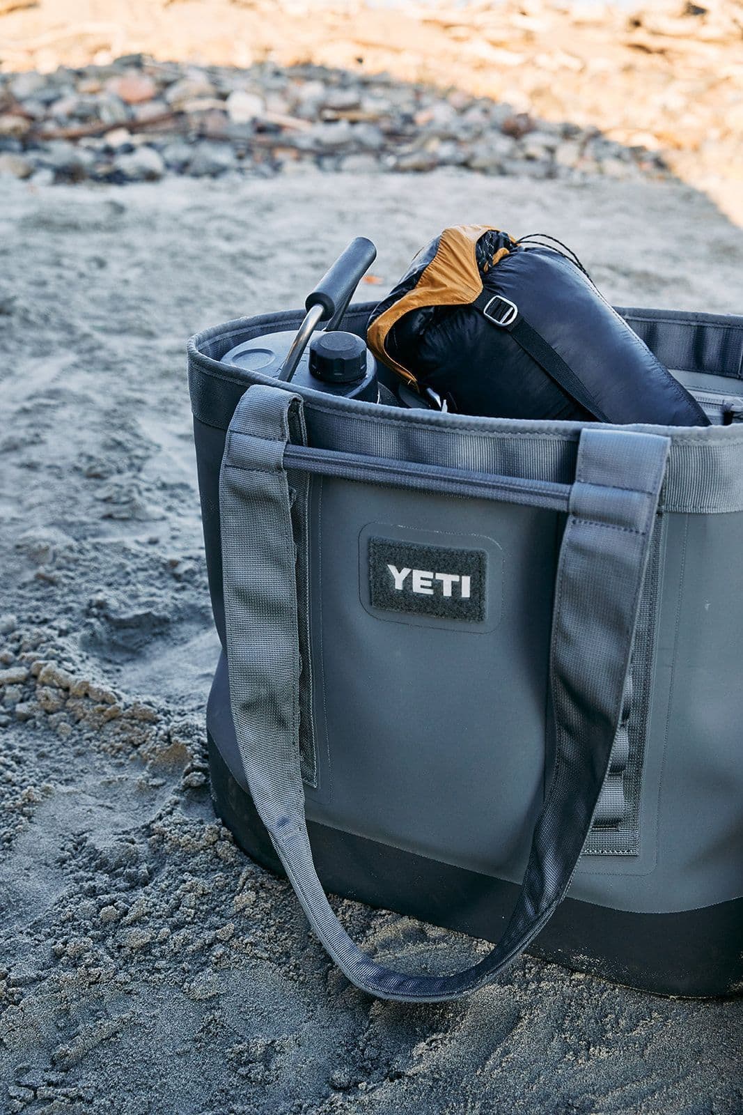 Eine YETI® Tragetasche Camino® Carryall 50 in Storm Grey sitzt auf sandigem Boden am Strand, gefüllt mit einer Wasserflasche, einem zusammengerollten Schlafsack und Outdoor-Ausrüstung. Im Hintergrund sind Felsen zu sehen.