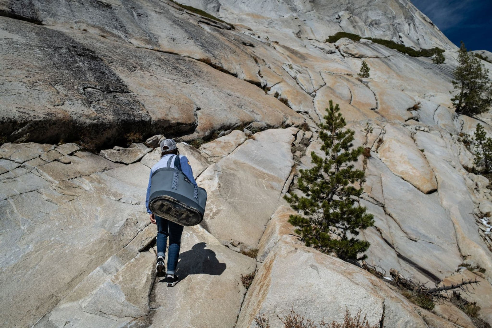Eine Person mit einer Isomatte und einer YETI® Tragetasche Camino® Carryall 50 - Storm Grey erklimmt einen steilen, felsigen Berghang mit spärlichen Bäumen und strukturiertem Granit unter einem klaren, wasserdichten Himmel.