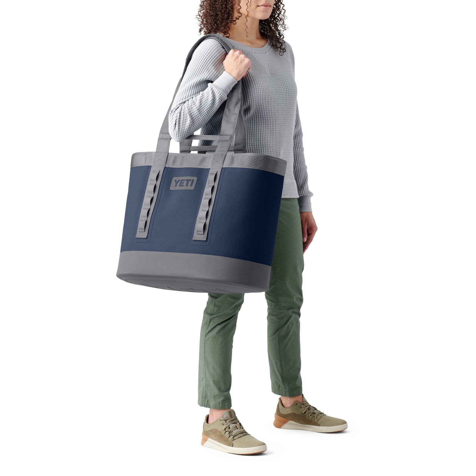 Eine Person, die einen grauen Pullover, eine grüne Hose und hellbraune Turnschuhe trägt, hat eine YETI® Tragetasche Camino® Carryall 50 - Storm Grey mit zwei stabilen Griffen und einem sichtbaren YETI-Logo dabei. Die wasserdichte Tasche wird über die Schulter gehängt.
