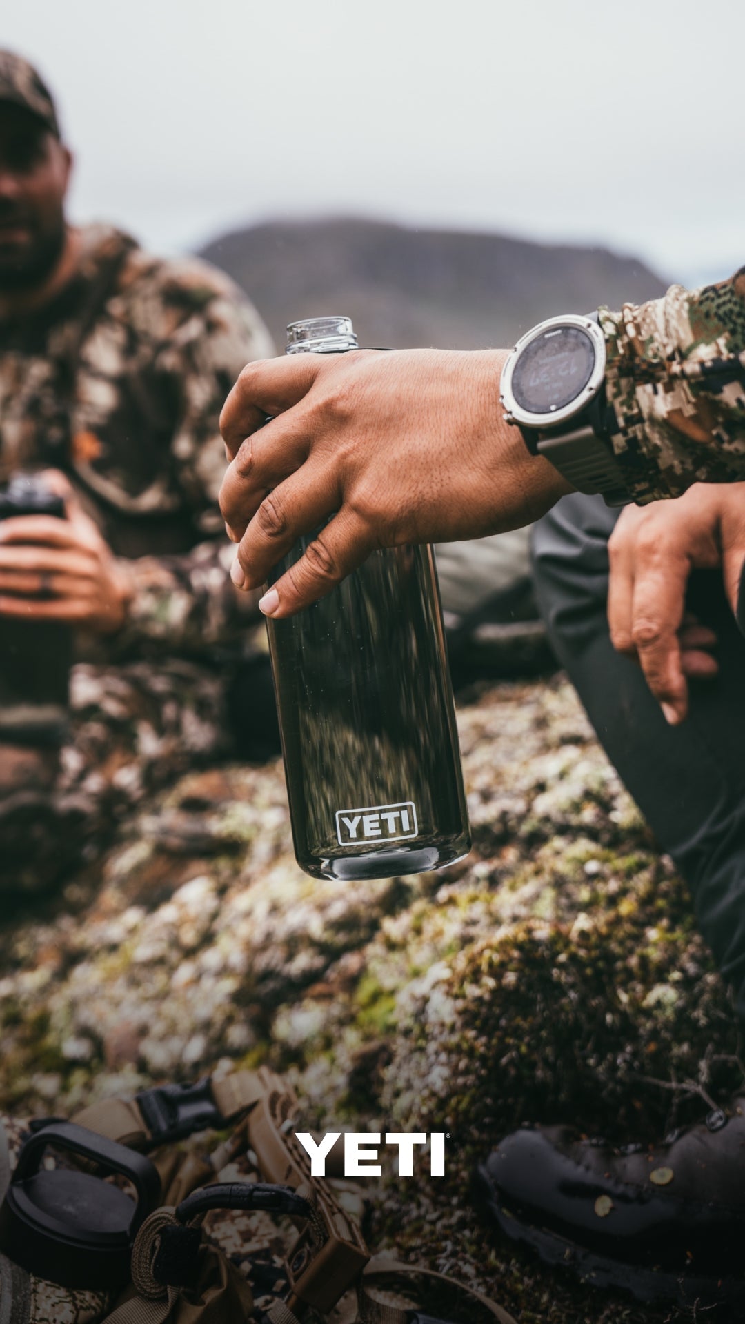 Eine Person in Tarnkleidung hält eine YETI® Yonder™ 750ml Wasserflasche - Rescue Red im Freien. Die auslaufsichere, BPA-freie Flasche sorgt für sichere Hydration. Das YETI Logo erscheint unten im Bild.