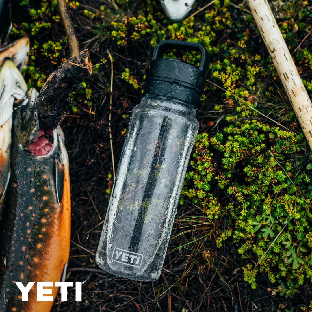 Eine klare YETI® Yonder™ 750ml Wasserflasche - Seafoam ruht auf grünem Moos neben einem frischen Fisch. Wassertropfen bedecken die BPA-freie Flasche, mit dem YETI-Logo und weißem Branding in der unteren linken Ecke.