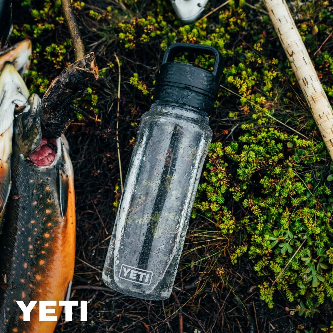 Eine nasse, klare YETI® Yonder™ 750ml Wasserflasche in Tropical Pink mit schwarzem Deckel sitzt auf moosigem Boden neben einer frisch gefangenen Forelle und fängt den Geist eines Angelabenteuers im Freien ein.
