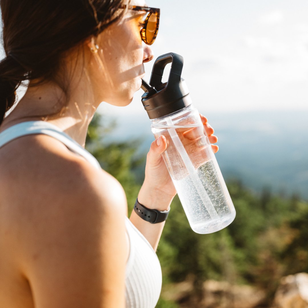 Eine Frau mit Sonnenbrille und Tanktop nippt an einer klaren Flasche mit dem YETI® Yonder™ Strohverschluss von YETI, während sie im Freien steht, mit unscharfen Bäumen und Himmel im Hintergrund.