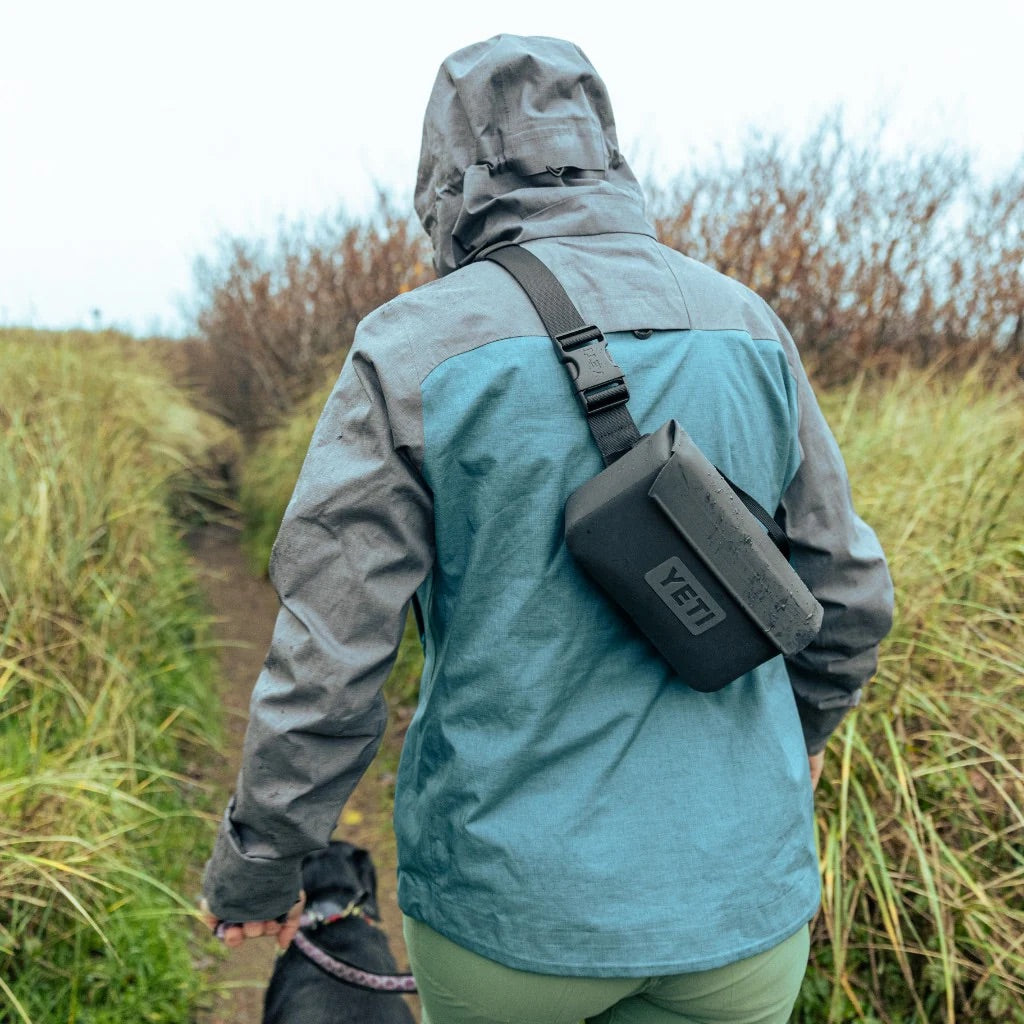 Eine Person in einer blau-grauen Regenjacke geht mit einem schwarzen Hund auf einem grasbewachsenen Weg spazieren und trägt die YETI® Zubehörtasche - Sidekick® 1L in Charcoal. Ihr Rücken ist der Kamera zugewandt, während der Himmel bedeckt ist.