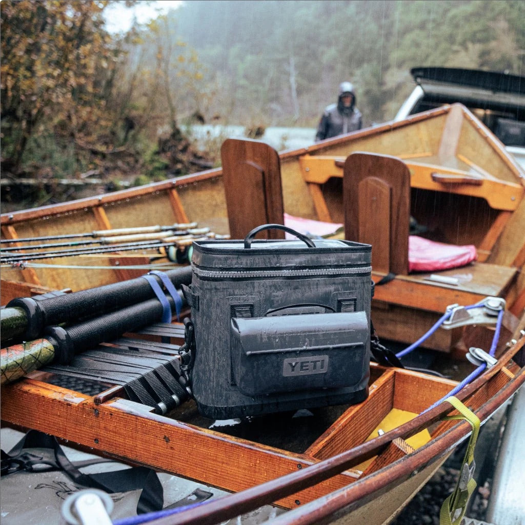 Nahaufnahme einer Angelausrüstung, einer YETI® Zubehörtasche - Sidekick® 1L in Charcoal und einer grauen YETI Kühlbox in einem Holzboot mit Angelruten; im nebligen Hintergrund erscheinen Bäume und eine Person.