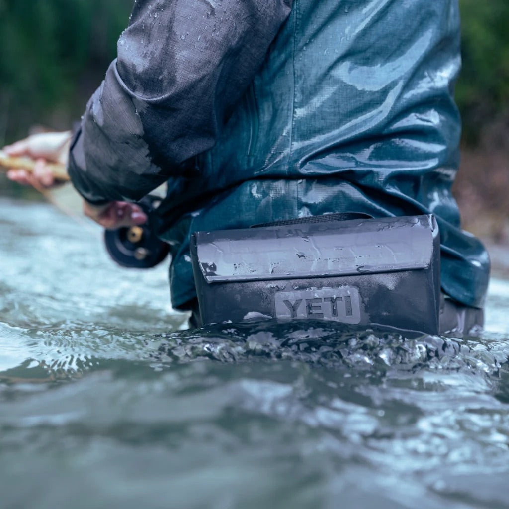 Eine Person in wasserdichter Ausrüstung angelt hüfttief in einem Fluss, das Wasser plätschert, während eine YETI® Zubehörtasche - Sidekick® 1L - Navy auf dem Rücken befestigt ist, während sie ihr Outdoor-Abenteuer erlebt.