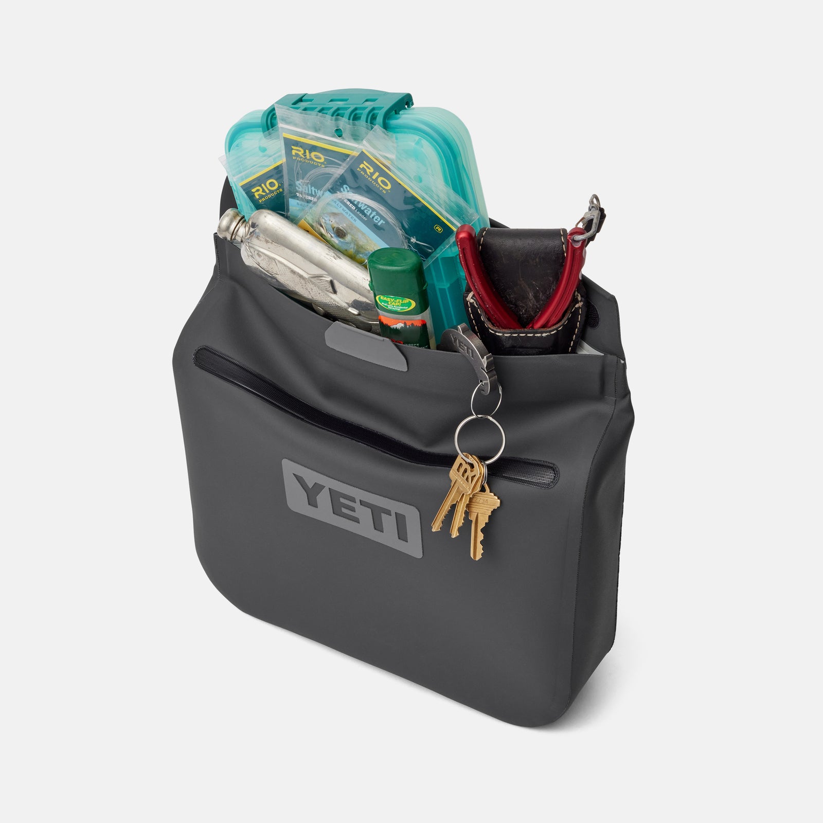 Eine YETI® Zubehörtasche - Sidekick® 6L in Charcoal bietet Platz für die Angelausrüstung: türkisfarbene Tacklebox, Multitool, rote Rolle, grüner Behälter. Ein Schlüsselbund hängt am Clip der vorderen Reißverschlusstasche. Marke: YETI.