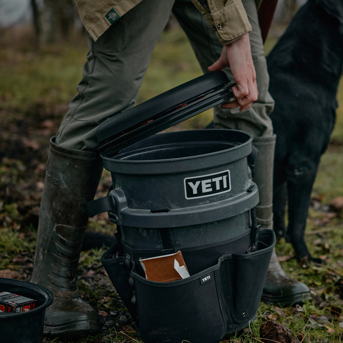 Eine Person in grünen Hosen und Stiefeln öffnet im Freien einen schwarzen YETI® drehbaren Eimerhocker. Der Eimer von YETI ist mit einem Deckel und Beuteln ausgestattet. In der Nähe steht ein schwarzer Hund auf Gras und Erde.